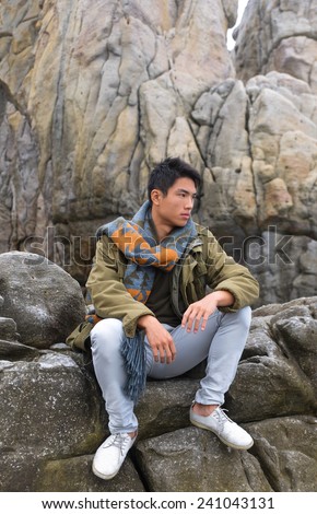 young man in jeans with scarf sitting on a rock at the sea watching the ocean