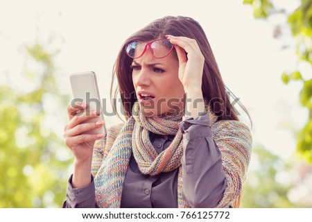Annoyed upset woman in glasses looking at her smart phone with frustration while walking on a street on an autumn day