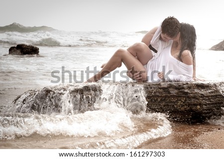 Young beautiful caucasian couple kissing on rock with wave breaking over them