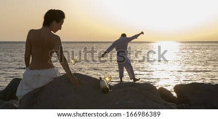 Young attractive topless caucasian woman watching man dance on beach rocks at sunrise
