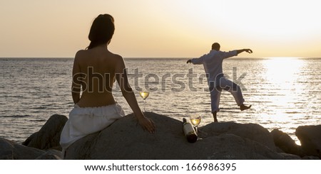 Young attractive topless caucasian woman watching man dance on beach rocks at sunrise