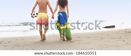 young couple walking along beach holding hands with football and Brazil flag