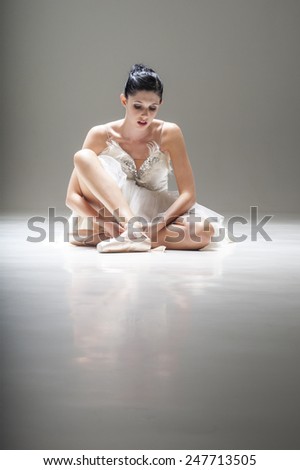 Young beautiful ballerina sitting under spotlight in costumed tutu adjusting pointed ballet shoe strapping