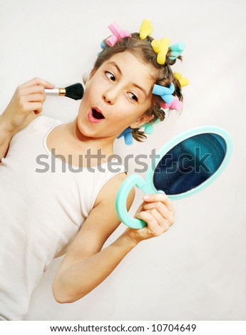 Young girl with colorful curlers, make-up brush and a mirror, pulling a funny face. More available.