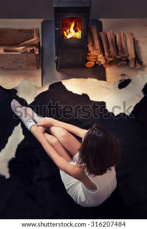 Beautiful young woman sitting down on a cow rug by the fireplace. Fall winter relaxing and leisure concept.