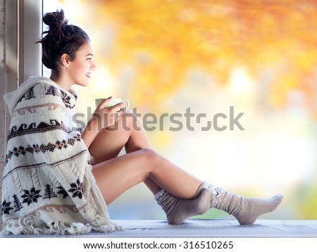 Young beautiful  brunette woman with cup of coffee wearing knitted nordic print poncho sitting home by the window. Blurred  garden fall background.