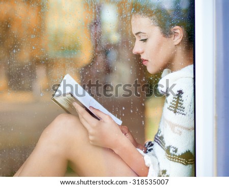 Beautiful young  brunette woman reading book wearing knitted dress sitting home behind a window covered with rain drops. Blurred fall garden reflection on the glass. Raining autumn concept