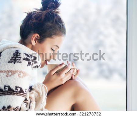 Young beautiful brunette woman drinking cup of coffee wearing knitted nordic print poncho sitting home by the window. Blurred winter snow tree background.