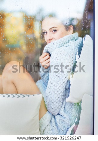 Beautiful young smiling brunette woman wearing knitted dress sitting home behind a window covered with rain drops. Blurred fall garden reflection on the glass