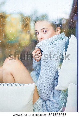 Beautiful young brunette woman wearing knitted dress sitting home behind a window covered with rain drops. Blurred fall garden reflection on the glass.
