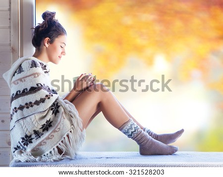 Young beautiful brunette smiling woman with cup of coffee wearing knitted nordic print poncho sitting home by the window. Blurred garden fall background.