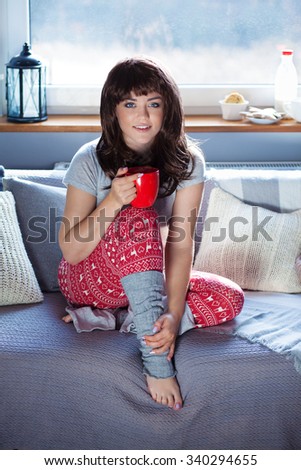 Young beautiful woman sitting home on the couch wearing Christmas print pajamas holding cup of coffee