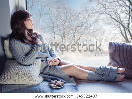Young beautiful brunette woman with cup of coffee and gingerbread wearing knitted cardigan sitting home relaxing by the window. Home chill out concept.