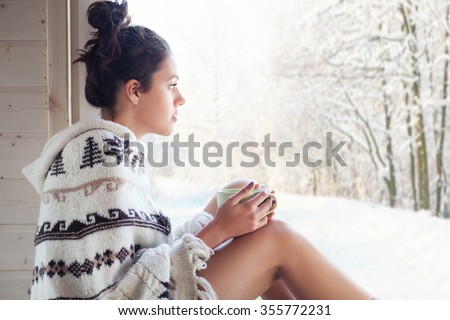 Young beautiful happy brunette woman drinking cup of coffee wearing knitted nordic print poncho chilling out home by the window. Winter forrest landscape background.