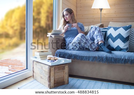Young beautiful blonde woman with cup of coffee and book sitting home by the window. Autumn fall trees view. Lazy day off concept
