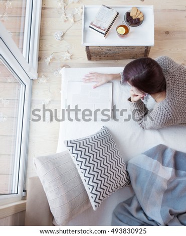 Woman lying down home on living room couch relaxing and reading magazine. Christmas lights on the floor, books and sweets on the table