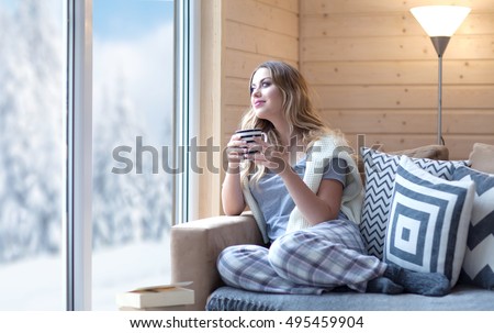 Young beautiful blonde woman with cup of coffee sitting home in living room by the window. Winter snow landscape view. Lazy day off concept