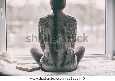 Thoughtful young brunette woman with book looking through the window, blurry winter forrest landscape outside