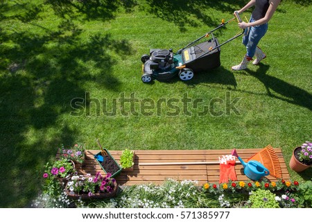 Woman wearing wellington boots mowing grass with lawn mower in the garden, gardening tools concept