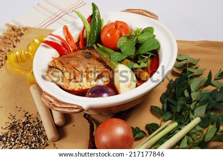 Bowl of spicy Asian curry fish head dish decorated with spices and ingredients on table mat.
