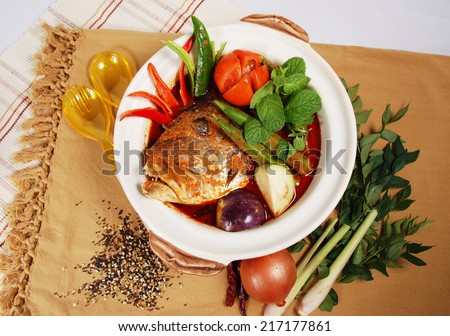 Bowl of spicy Asian curry fish head dish decorated with spices and ingredients on table mat.