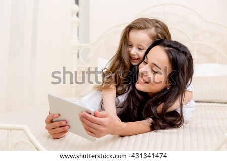 beautiful young mother and her cute daughter in white shirts and jeans lying on the bed at home , laughing and looking in digital tablet