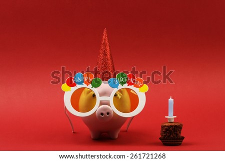 Piggy bank with sunglasses Happy birthday, party hat and birthday cake with blue candle on red background