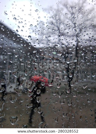 Photo of a street, building, person with red umbrella and tree on a rainy day trough a wet window with a lot of water drops all over it