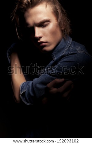 Handsome young man in jean jacket with beautiful face, fashionable hairstyle and crossed hands looking down