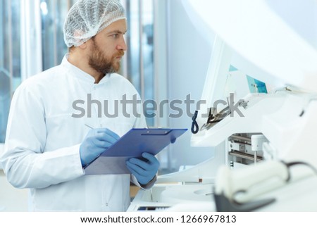 Handsome laboratory assistant in white cap and blue gloves doing experiments. Attractive doctor with red beard in lab coat checking new modern equipment. Young medical worker writing results.