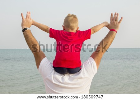 son in a red shirt sitting on his father\'s shoulders and looks at the sea