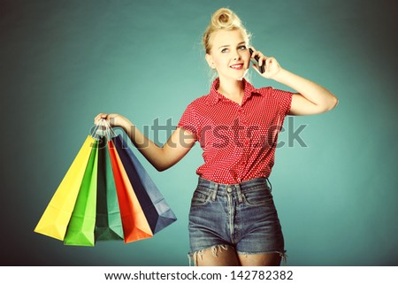 Retro style. Young woman in red dotted shirt and shorts with colourful shopping bags talking on cell phone on blue background