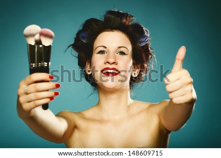 young woman preparing to party having fun, girl styling hair with curlers and make up paint brushes applying make up retro style blue background