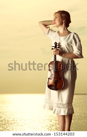 The blonde girl music lover on pier with a violin at sunset or sunrise.  Love of music concept.