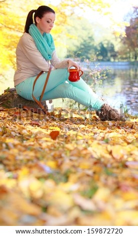 Young thoughtful pensive woman in the autumn park relaxing enjoying hot drink coffee or tea, holding red mug with warm beverage. Yellow leaves background