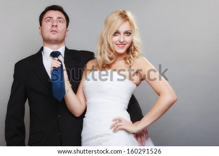 Wedding day. Portrait of happy married couple blonde bride and groom. Woman pulling on mans tie, studio shot on gray background