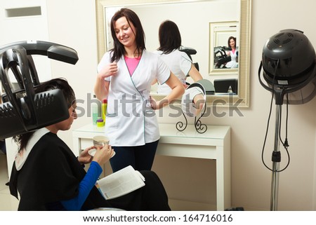 Young woman female client with magazine drinking coffee tea in hairdressing beauty salon. Girl dying colouring hair by hairdresser hairstylist. Modern equipment.