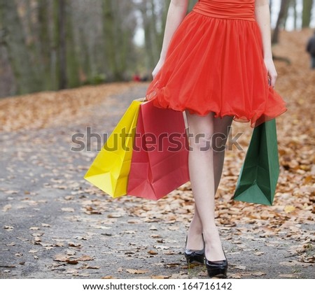 Close up of female legs. Girl in elegant red dress with colorful paper shopping bags walking in the autumnal park. Sale and retail.