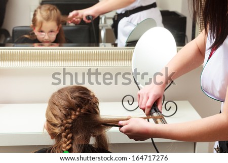 Reflection on mirror of kid by hairstylist. Hairdresser with straightener straightening combing hair of little girl in eyeglasses. Child in hairdressing beauty salon.
