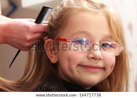 Happy smiling kid by hairstylist. Hairdresser with comb combing hair of little girl in eyeglasses. Child in hairdressing beauty salon.