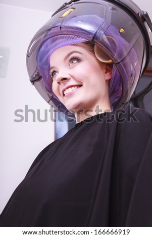 Smiling young woman female client in hairdressing beauty salon. Girl in hair rollers curlers with hairdryer dryer relaxing by hairdresser hairstylist.
