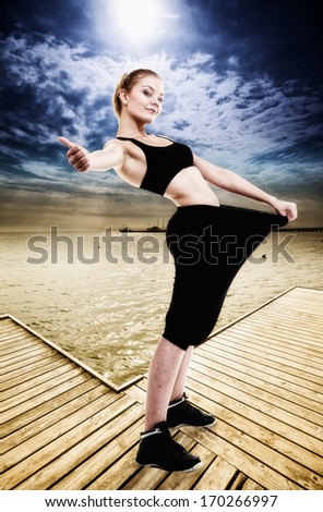 Weight loss and healthy lifestyle. Happy woman fitness girl showing how much weight she lost, giving thumb up success hand sign. Outdoor. Beach pier