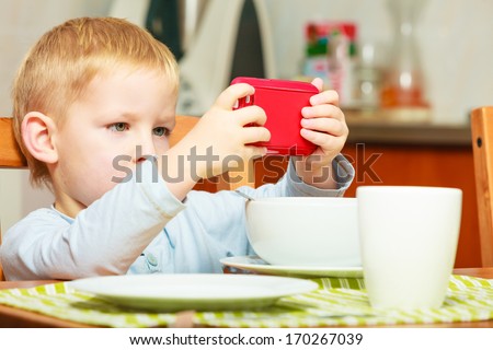 Happy childhood. Blond boy kid child eating corn flakes cereal with milk breakfast morning meal playing with mobile phone at the table. Home.