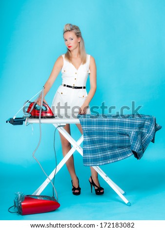Full length sexy girl retro style ironing male shirt, woman housewife in domestic role. Traditional sharing household chores.  Pin up housework.  Vivid blue background
