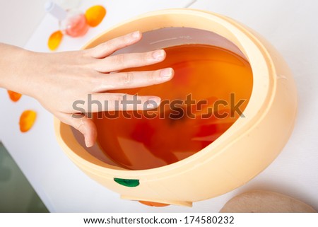Female hand and orange paraffin wax in bowl. Manicure and skincare. Woman girl in beauty spa salon.