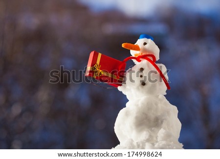 Little happy snowman in blue screw top as hat red scarf and with fork christmas gift box outdoor. Winter season seasonal specific. Blurred background.
