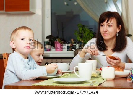 Family mother and two blond brothers boys kids children eating corn flakes and bread breakfast morning meal at the table. Home.