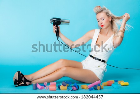 Full length young woman preparing to party, sexy girl fashion model styling hair with curlers accessories and hairdryer retro style vivid blue background