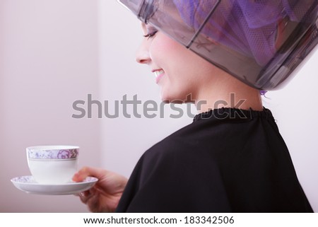 Young woman female client drinking hot beverage coffee tea in hairdressing beauty salon. Girl in hair rollers curlers with hairdryer dryer relaxing by hairdresser hairstylist.
