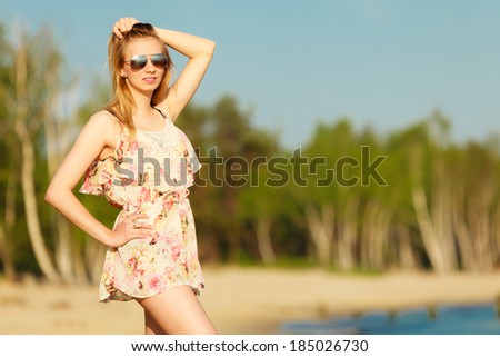 Vacation. Girl in summer dress and sunglasses standing on the empty beach. Young woman relaxing on the sea coast. Summertime.